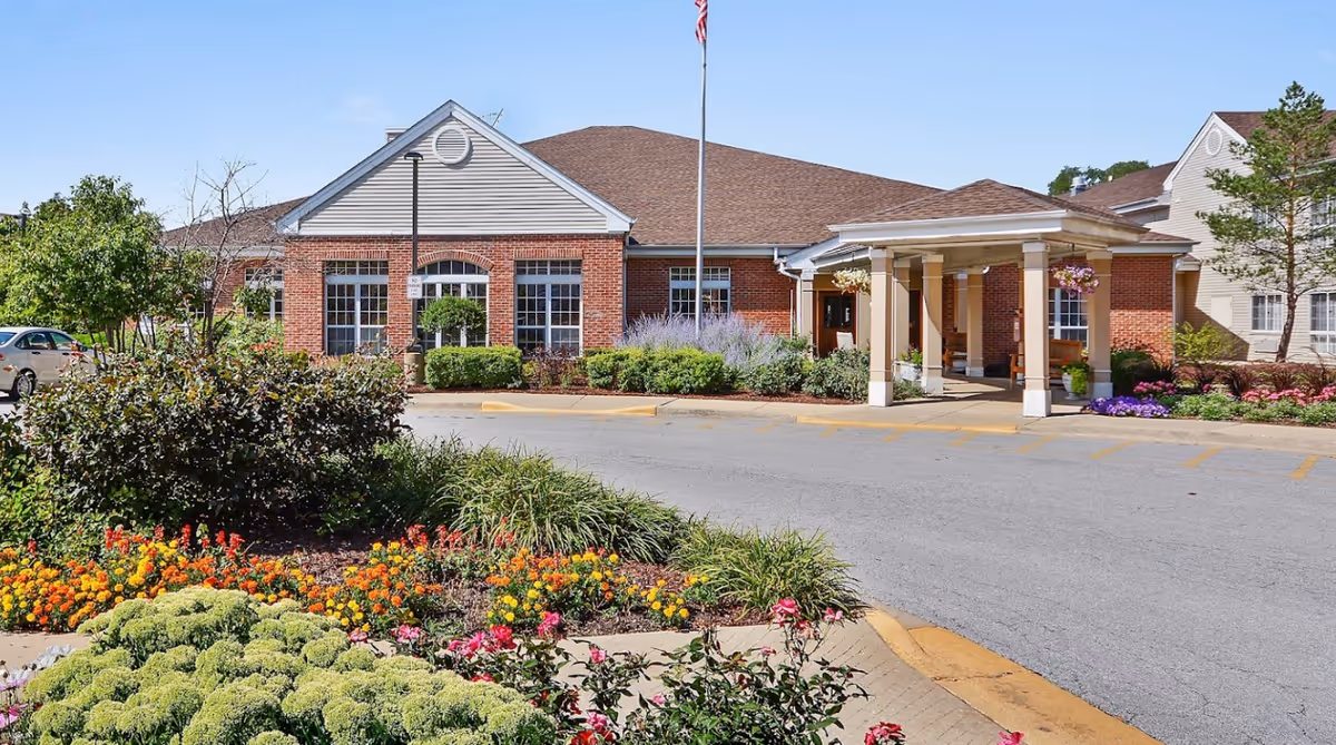 Exterior view of Eden Vista Prospect Heights facility showing a brick building with large windows, a covered entrance with columns, an American flag on a flagpole, and landscaped flower beds with colorful flowers and greenery in the foreground under a clear blue sky.