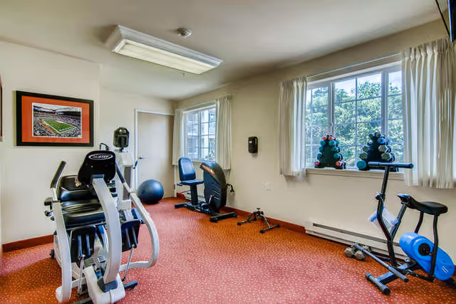 A small fitness room with exercise equipment including a recumbent bike, an elliptical machine, a stationary bike, and a set of dumbbells on a rack by the window. The room has beige walls, a red carpeted floor, and two windows with white curtains letting in natural light.