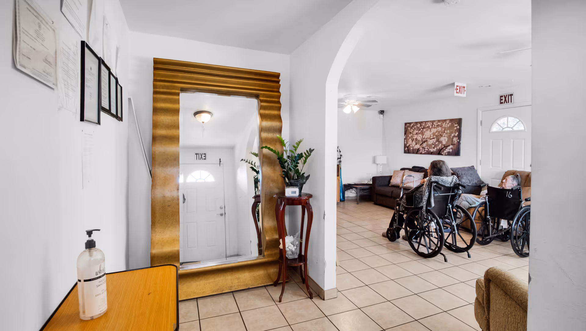 Interior view of a senior living facility common area with two elderly individuals in wheelchairs facing a brown couch and a wall-mounted artwork. The room has tiled floors, white walls, and two exit signs above doors. A large gold-framed mirror and a small table with a plant and hand sanitizer are visible in the foreground.