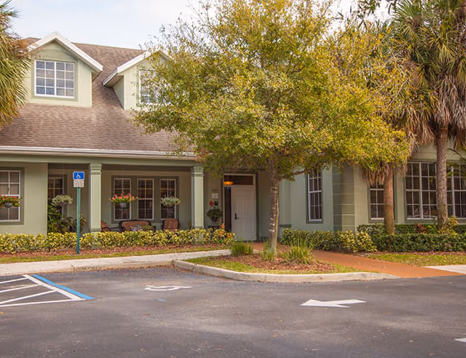 Exterior view of a senior living facility building with a green facade, multiple windows, a covered porch with chairs, a tree in front, and a parking area with a handicap parking space.
