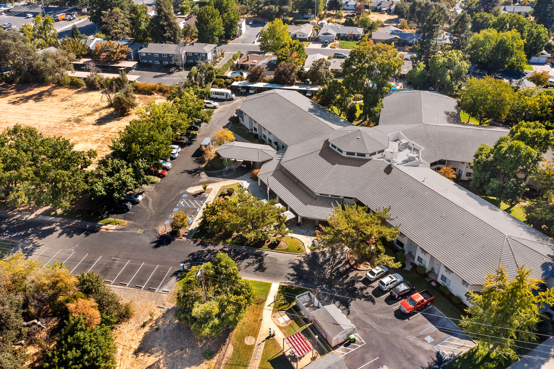 Aerial view of Provincial Chico Senior Living facility showing a large building with a gray roof surrounded by trees, parking lots with several cars, and nearby residential houses.