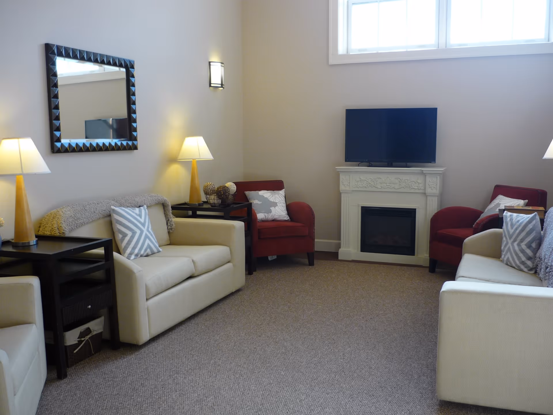 A cozy living room with two beige sofas and two red armchairs arranged around a white decorative fireplace with a flat-screen TV on top. The room has beige walls, a large window near the ceiling, two table lamps on black side tables, a wall-mounted light fixture, and a large framed mirror on one wall.