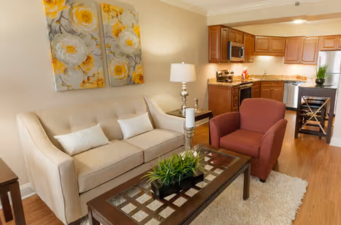 A cozy living room area with a beige sofa adorned with two white pillows, a maroon armchair, and a glass-top coffee table with a green plant and candle holders. Behind the living area is a kitchen with wooden cabinets, stainless steel appliances, and a small dining area with a high chair. The walls are decorated with two large floral paintings featuring yellow and white flowers.