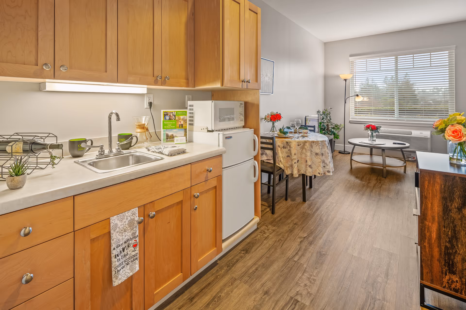 A bright and cozy kitchen and dining area in a senior living facility. The kitchen features wooden cabinets, a sink, a small refrigerator, and a microwave. On the counter, there are mugs, a wine bottle holder, and a small plant. The dining area has a round table covered with a floral tablecloth, set with plates, glasses, and a vase with red flowers. A window with blinds lets in natural light, and there is a floor lamp and some greenery near the window. The floor is wooden, and the overall atmosphere is warm and inviting.
