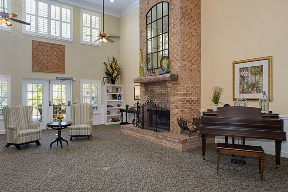 A spacious living room area with two striped armchairs and a small round table with flowers between them. There is a large brick fireplace with a decorative black metal screen and a tall mirror above the mantel. To the right, there is a dark wooden piano with a bench, a framed painting on the wall, and some decorative items on top of the piano. The room has high ceilings with ceiling fans and large windows letting in natural light. A built-in white bookshelf with various decorative items is next to the fireplace.