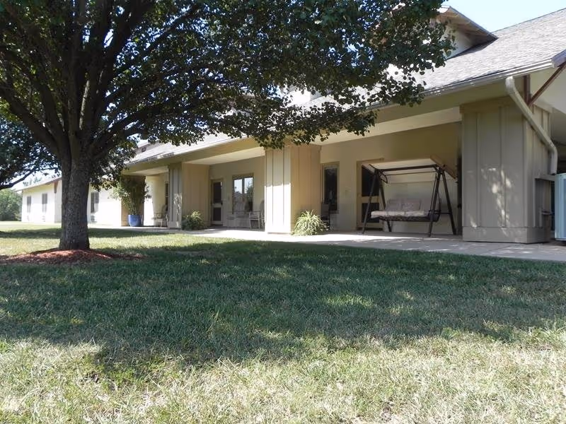 Exterior view of a single-story building with a covered porch, outdoor swing, and a large tree on a grassy lawn.