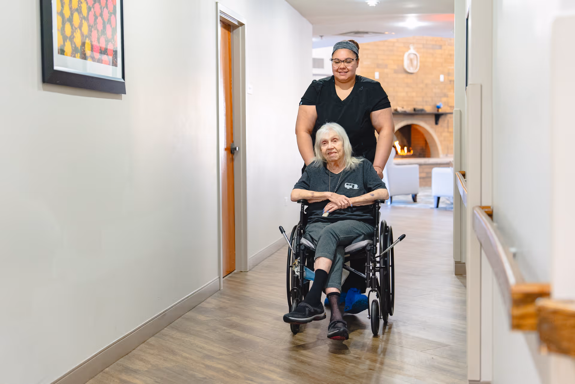 A caregiver is pushing an elderly woman in a wheelchair down a hallway inside a supportive living facility. The hallway has light-colored walls, wooden flooring, handrails on the right side, and a framed artwork on the left wall. In the background, there is a cozy seating area with a fireplace and chairs.