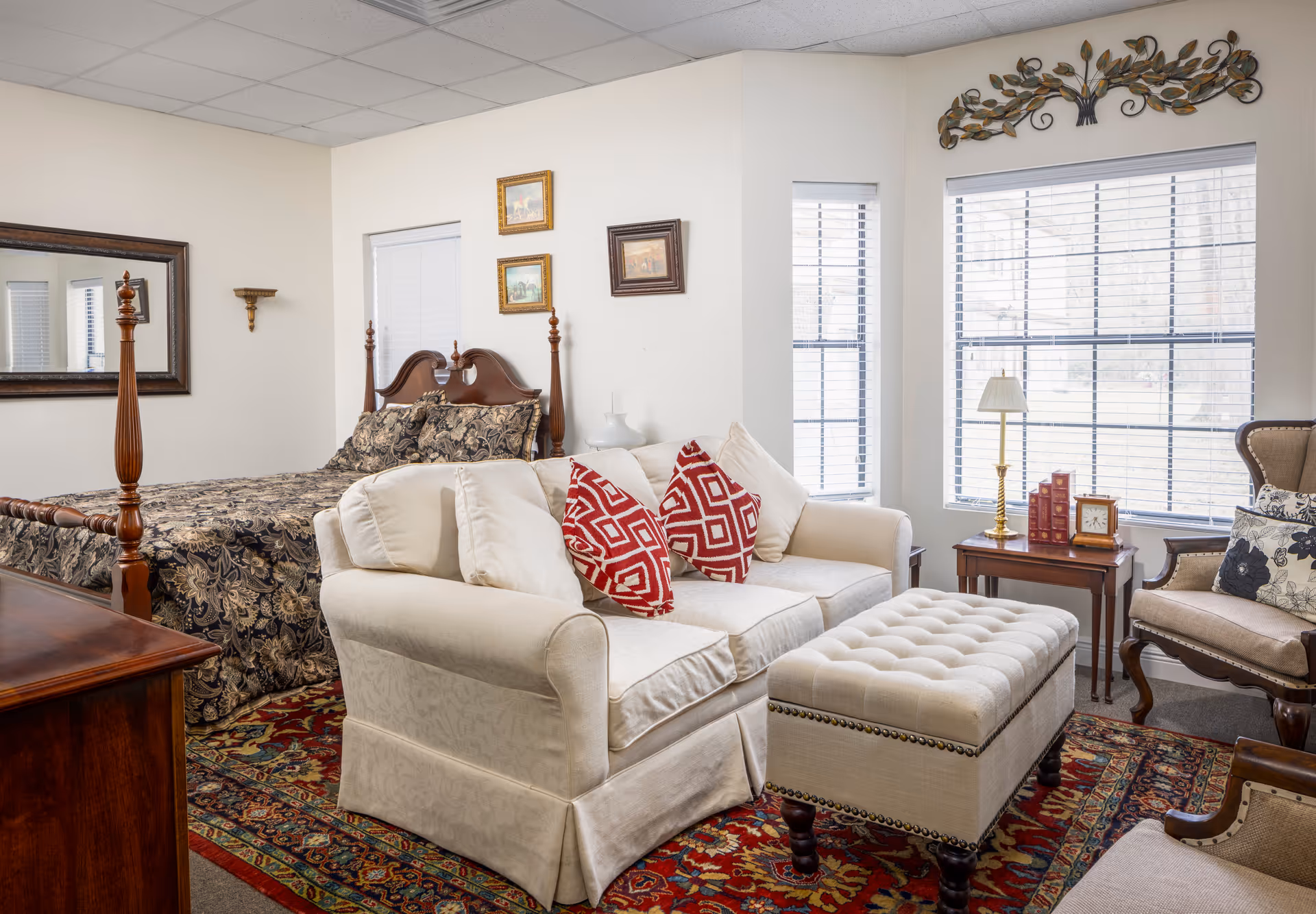 A cozy bedroom and sitting area in an assisted living facility featuring a wooden four-poster bed with patterned bedding, a cream-colored loveseat with red and white patterned pillows, a tufted ottoman, an armchair with floral cushions, a wooden side table with a lamp, books, and a clock, and large windows with blinds letting in natural light.