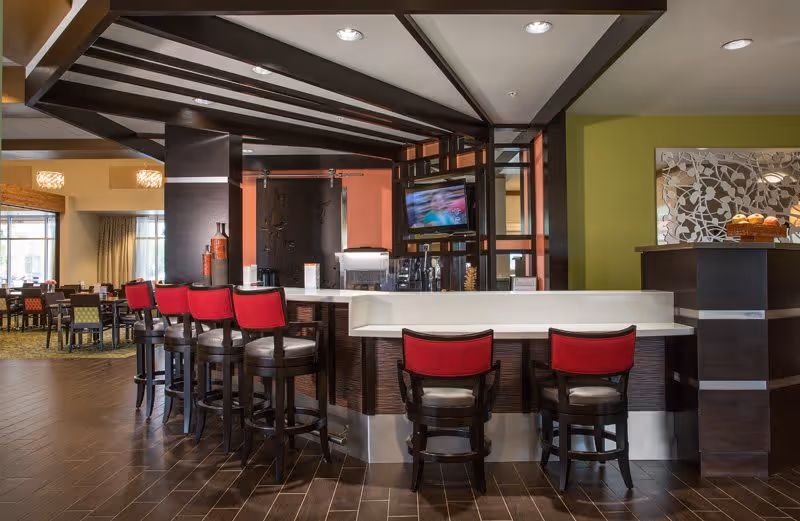 Interior view of a modern dining area with a bar counter featuring six high chairs with red backs. The space has wooden flooring, a green accent wall, a mounted TV, and additional dining tables and chairs in the background near large windows.