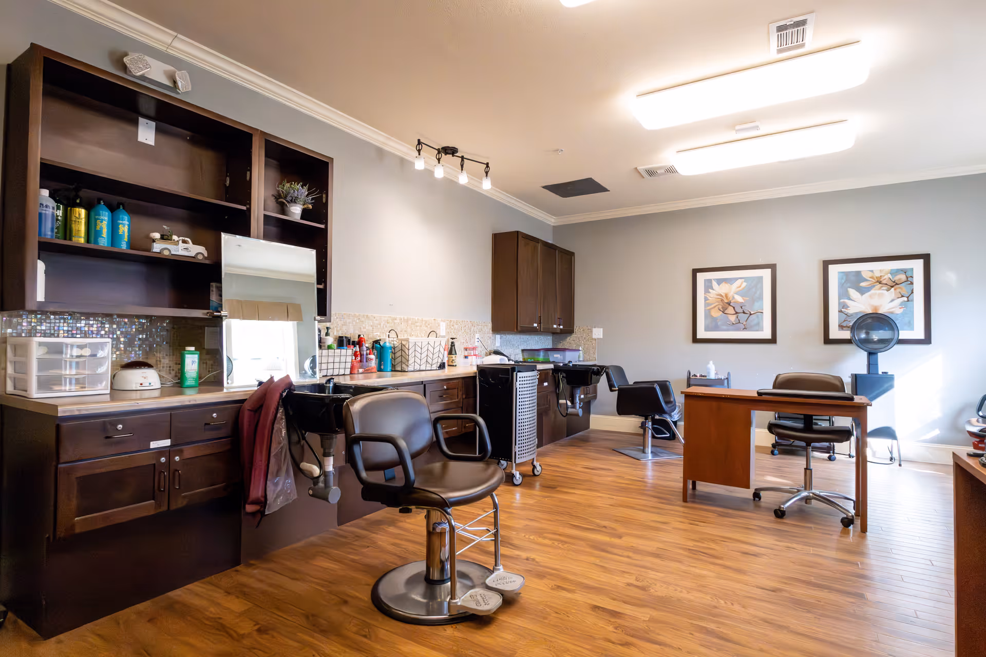 Interior salon room with styling chairs, counters, a shampoo sink and desks on wood flooring.