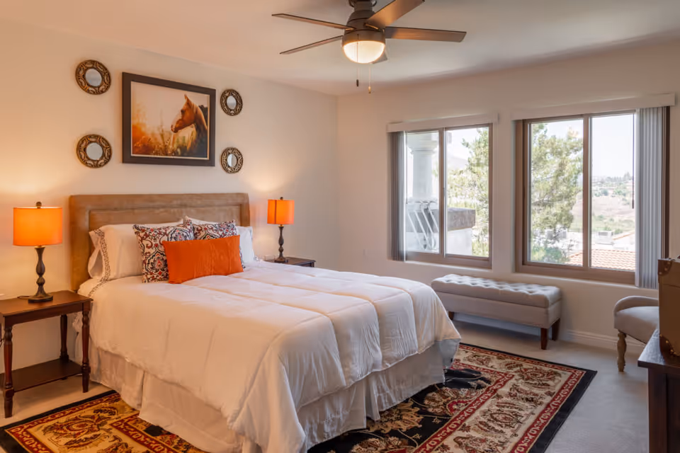 A cozy bedroom with a large bed featuring white bedding and decorative pillows. Two bedside tables each hold a lamp with an orange shade. Above the bed is a framed picture of a horse surrounded by four circular decorative mirrors. The room has a ceiling fan with a light, two large windows with vertical blinds, a cushioned bench under the windows, and a patterned area rug on the floor.