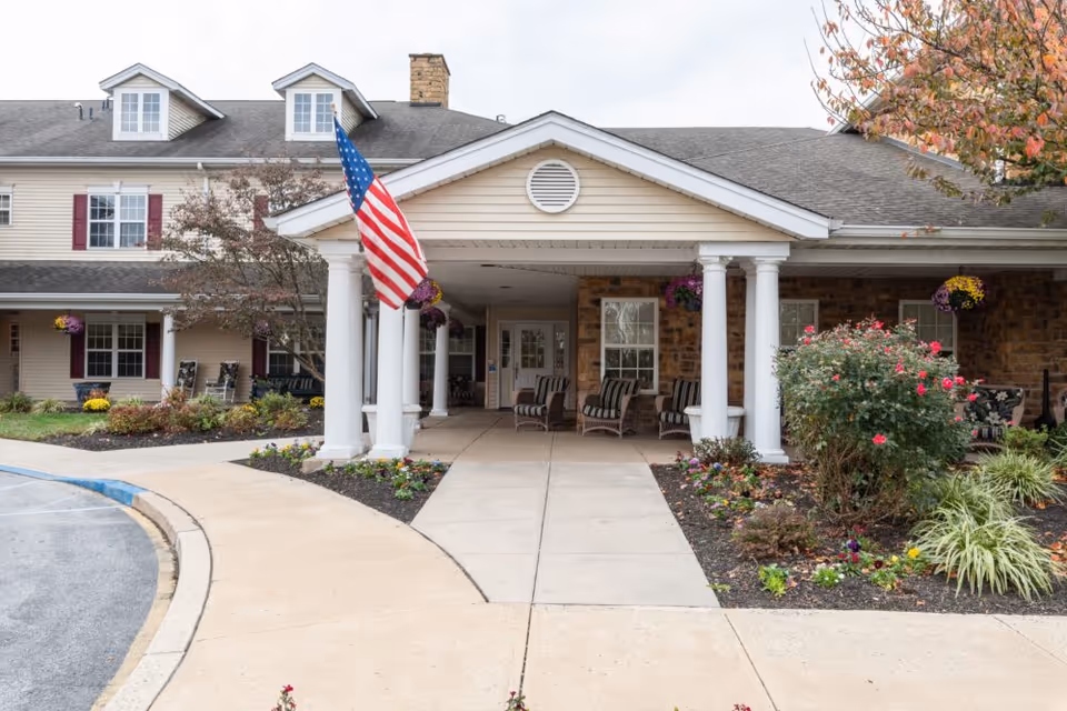Entrance of Somerford House And Place Newark showing a covered porch with white columns, an American flag, outdoor seating with striped cushions, and landscaped flower beds with bushes and hanging flower baskets.