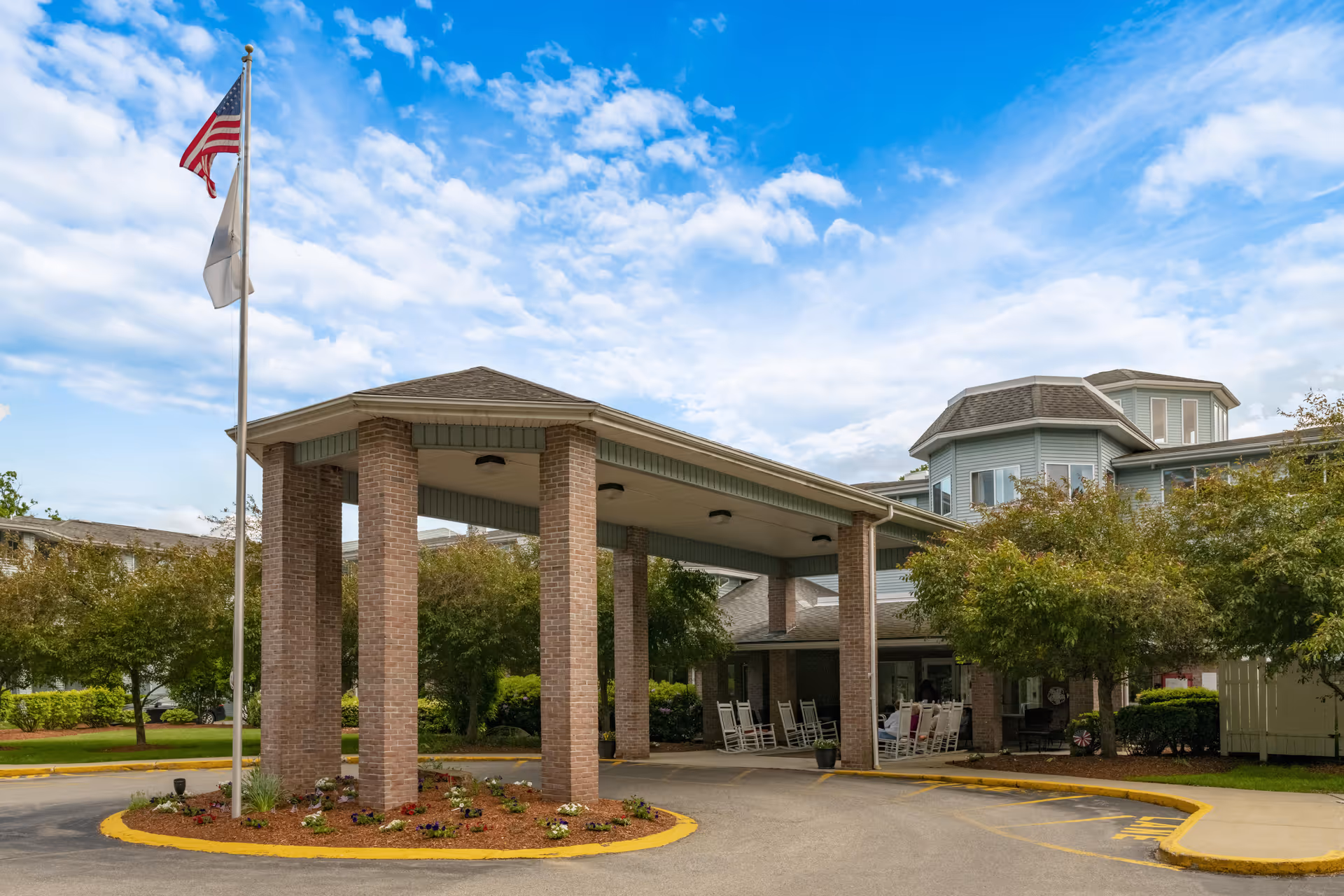 Entrance of Summer Place - A Provincial Senior Living Community featuring a covered drop-off area supported by brick pillars, an American flag on a flagpole, several rocking chairs under the covered area, and surrounding greenery with trees and shrubs under a partly cloudy blue sky.