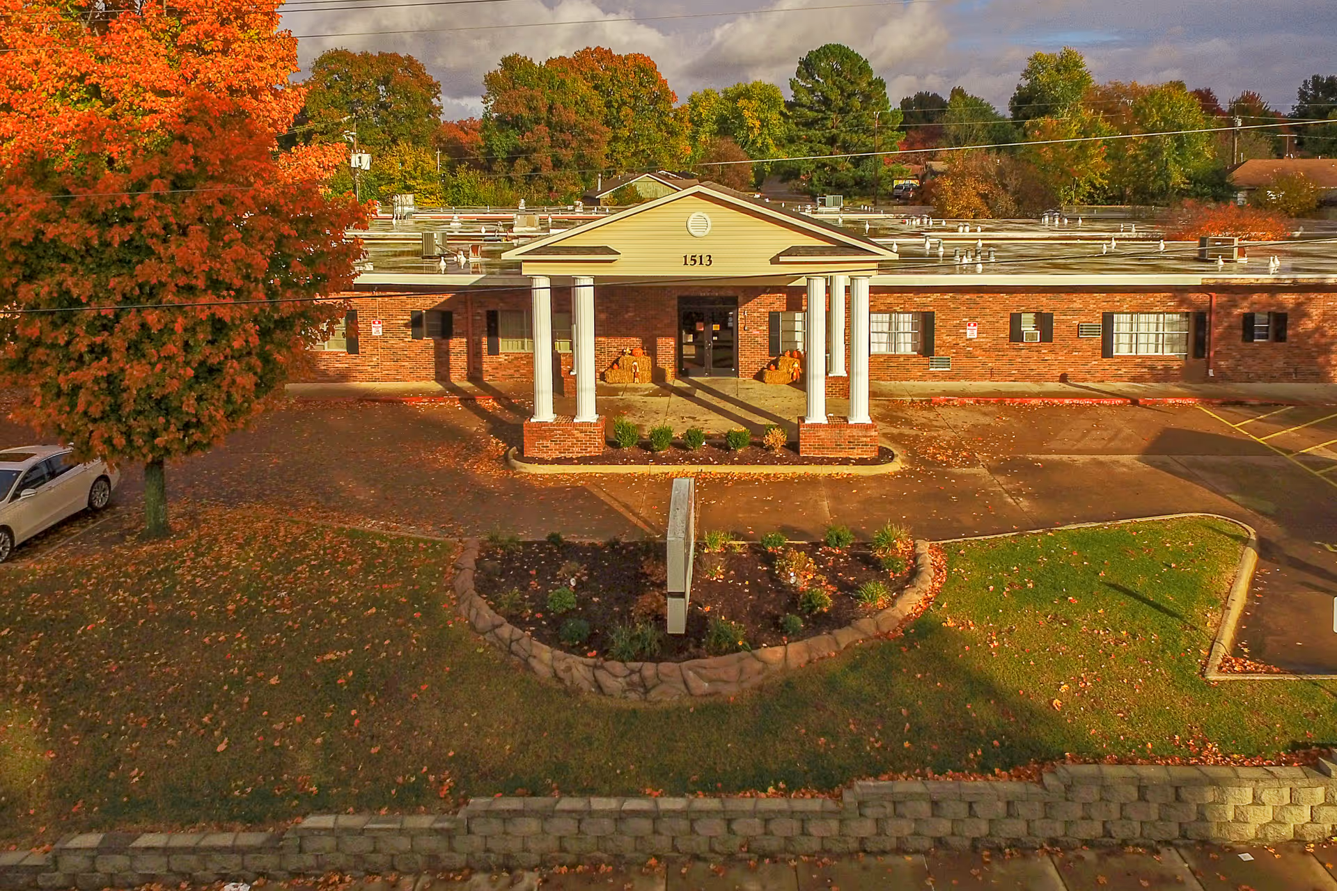 Front exterior view of a single-story brick building with white columns at the entrance, surrounded by autumn trees with orange and green leaves. The building has the number 1513 above the entrance and a small garden area in front with a stone border.