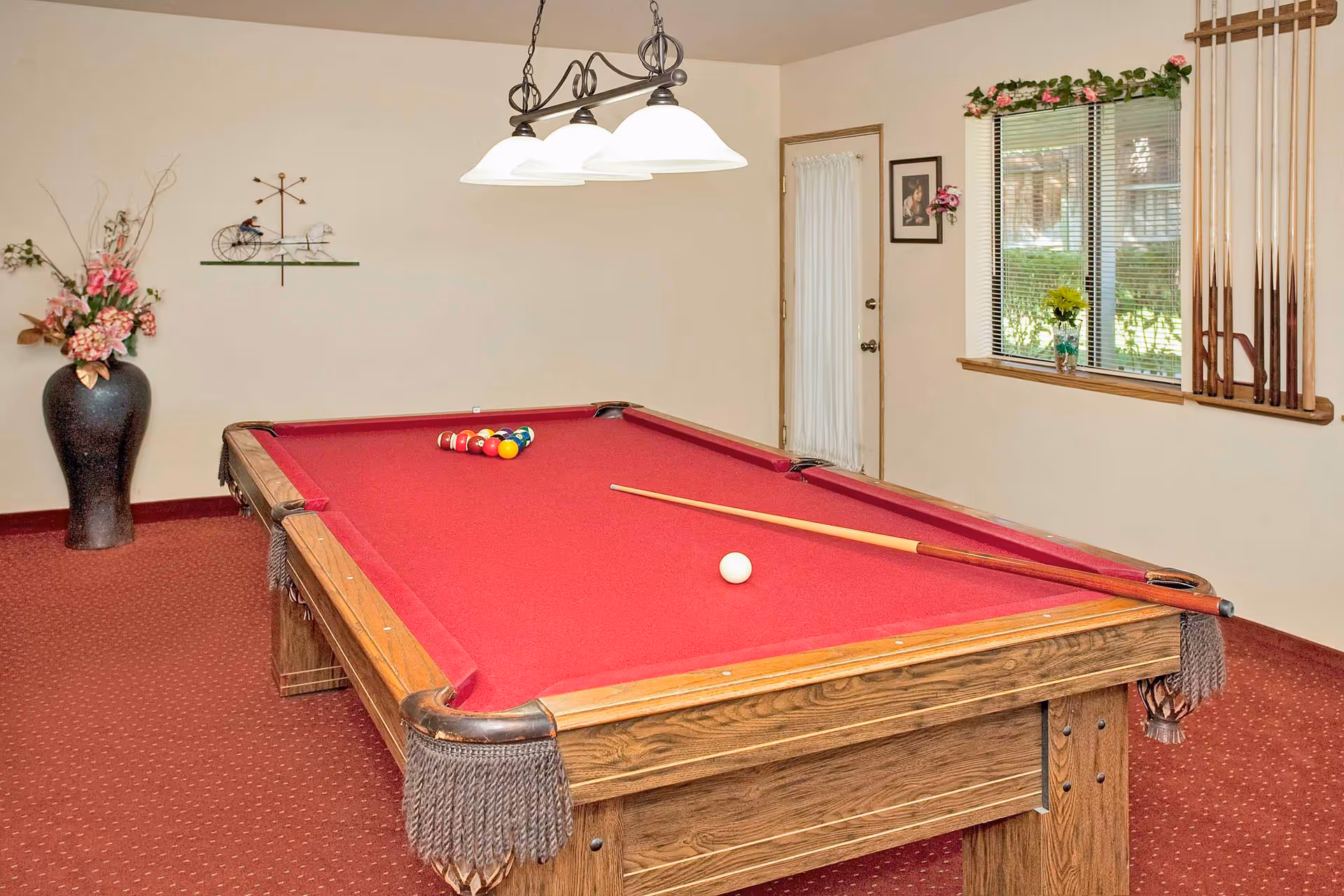 Indoor recreation room featuring a red-felt pool table with cues, overhead lights, and a window with a cue rack on the wall.