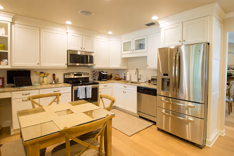 Bright and modern kitchen with white cabinets, stainless steel appliances including a refrigerator, oven, microwave, and dishwasher. A wooden dining table with four chairs and placemats is in the foreground. The kitchen has a light wood floor and recessed lighting.