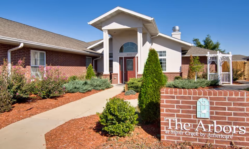 Exterior view of a single-story brick building with a peaked roof and a covered entrance. There is a concrete walkway leading to the red front door, surrounded by landscaped bushes and trees. A brick sign in the foreground reads 'The Arbors at Silver Creek by Americare' with a decorative window logo above the text. The sky is clear and blue.
