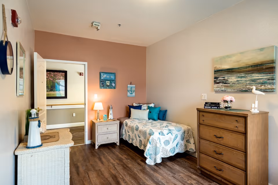 A cozy bedroom in a senior living facility with a single bed covered in a seashell-patterned quilt and multiple blue and white pillows. Next to the bed is a small nightstand with a lamp and decorative lighthouse. Across from the bed is a wooden dresser with a vase of pink flowers, books, and a bird figurine on top. The walls are painted in soft beige and light brown tones, with nautical-themed artwork hanging above the bed. The room has wood flooring and an open door leading to a hallway with a framed picture on the wall.