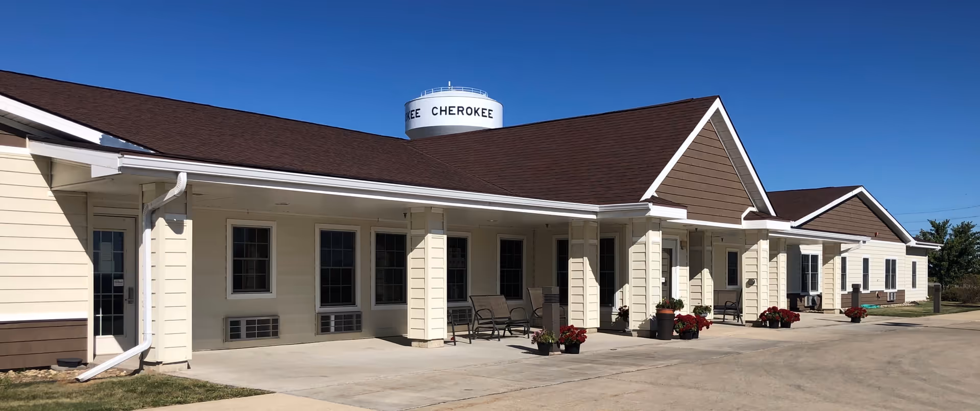 Exterior view of a single-story building with beige siding and a brown roof under a clear blue sky. The building has multiple windows and a covered walkway with several benches and potted plants. A water tower labeled 'CHEROKEE' is visible in the background.