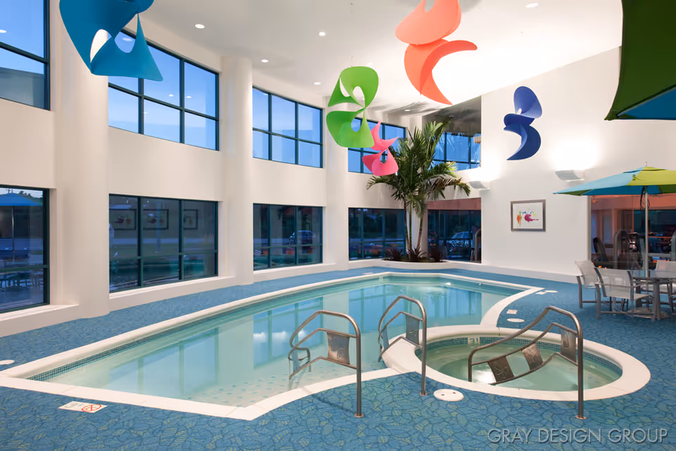 Indoor swimming pool area with large windows, colorful hanging decorations, a hot tub, and seating with tables and umbrellas.