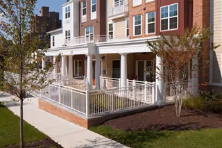 Exterior view of a multi-story senior living facility building with brick and light-colored siding, featuring a covered porch with white railings and columns, surrounded by landscaped greenery and a sidewalk.