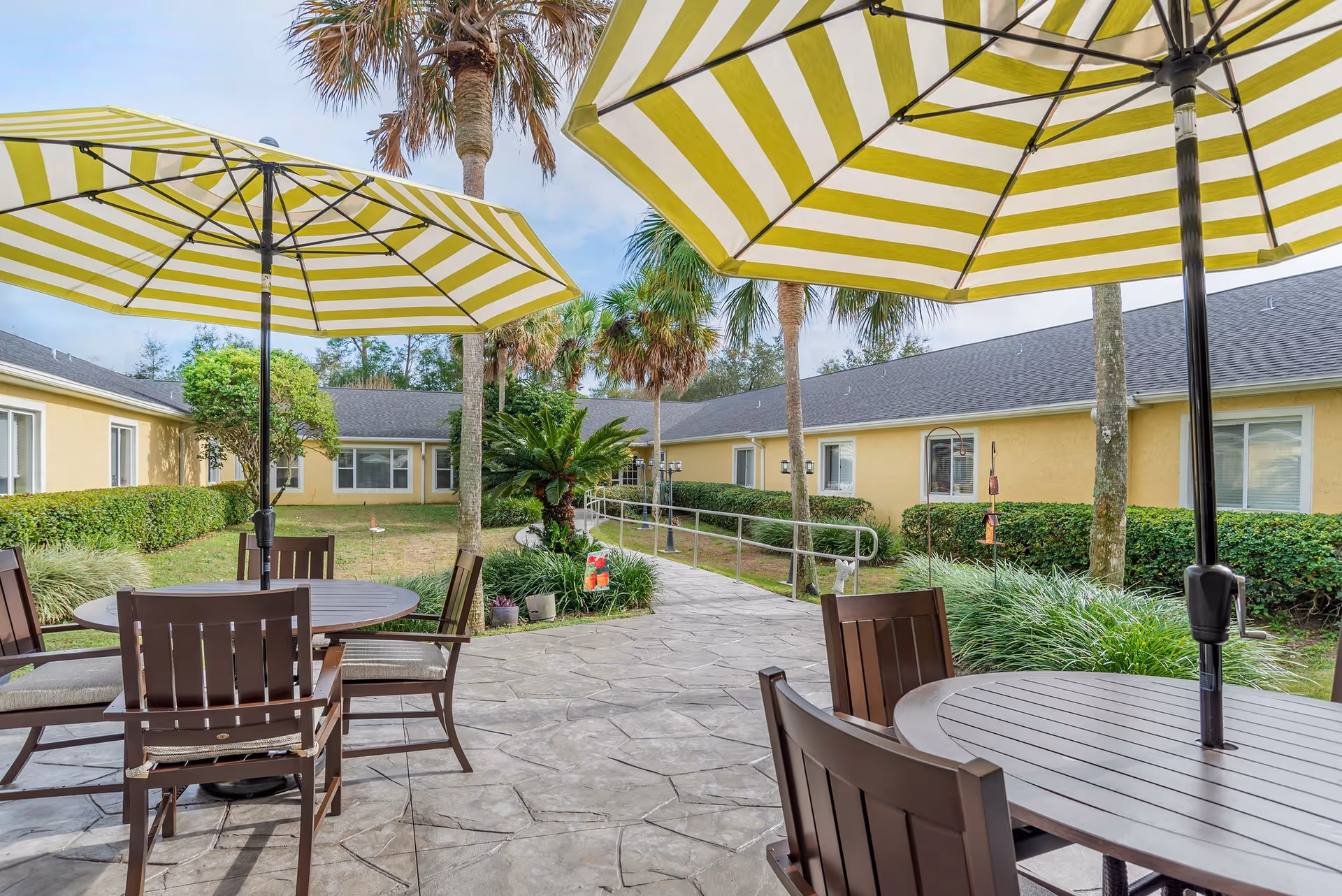 Outdoor patio area with round tables and brown chairs under large yellow and white striped umbrellas. The patio is surrounded by a garden with palm trees, bushes, and a paved walkway leading to a yellow building with multiple windows.