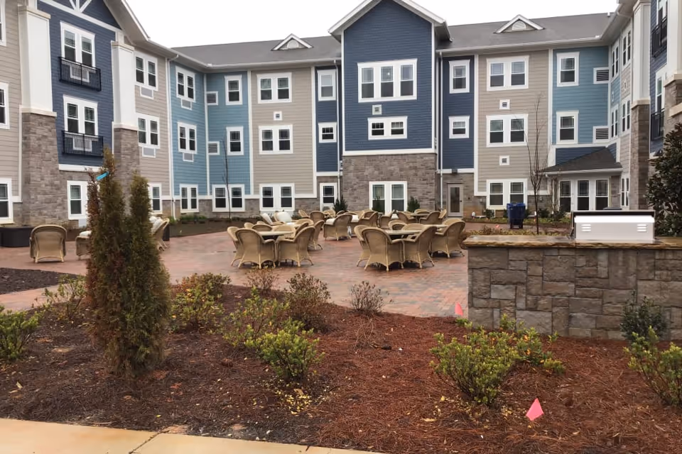 Outdoor courtyard area of a senior living facility with multiple round tables and wicker chairs arranged on a brick patio. The surrounding building has three stories with blue, beige, and gray siding and white-trimmed windows. There are small landscaped garden beds with shrubs and mulch in the foreground.