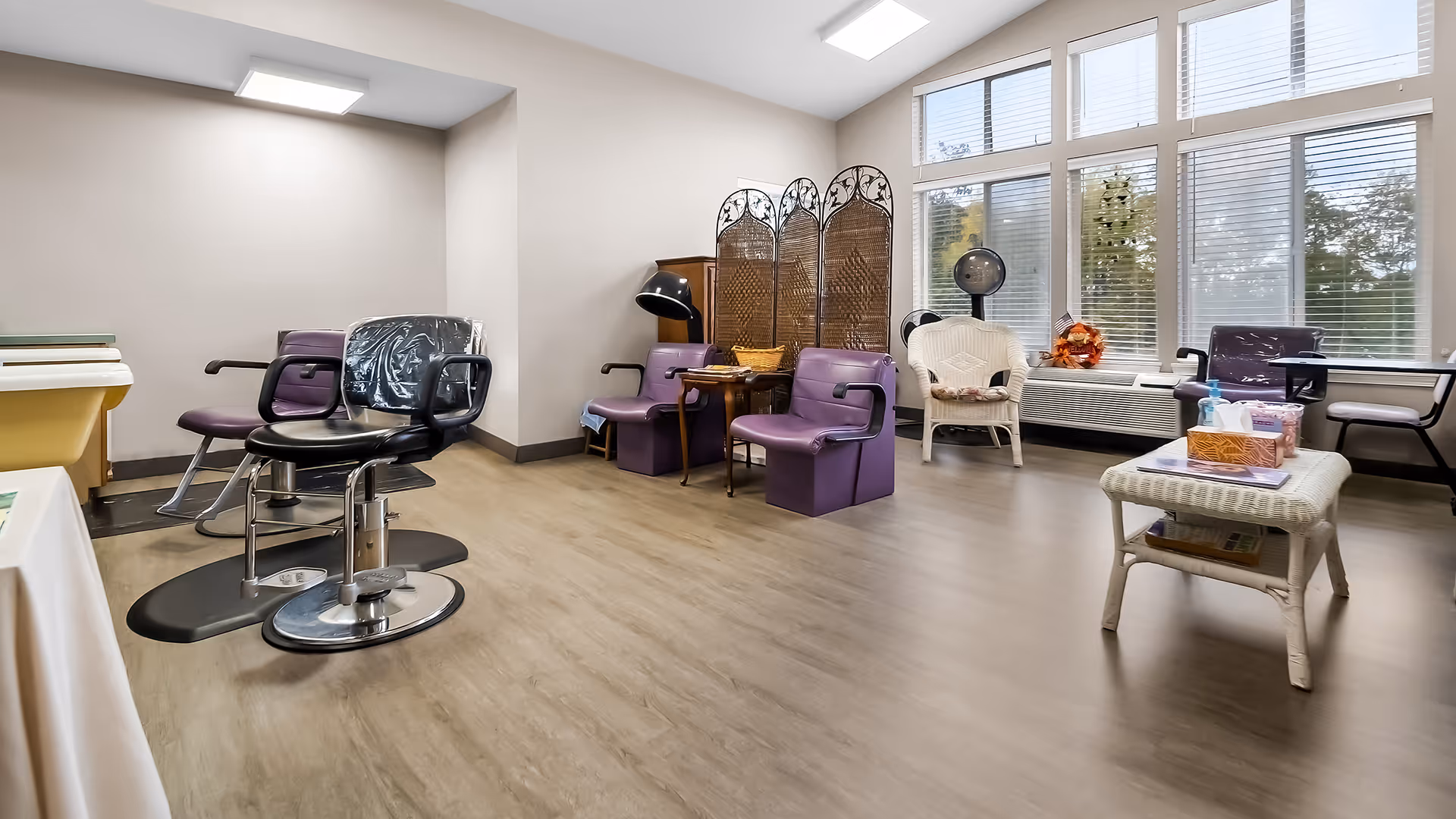 Interior view of a salon area in a senior living facility with several purple salon chairs, a black salon chair, a wicker chair, a wooden folding screen, and a table with tissues and hand sanitizer near large windows with blinds.