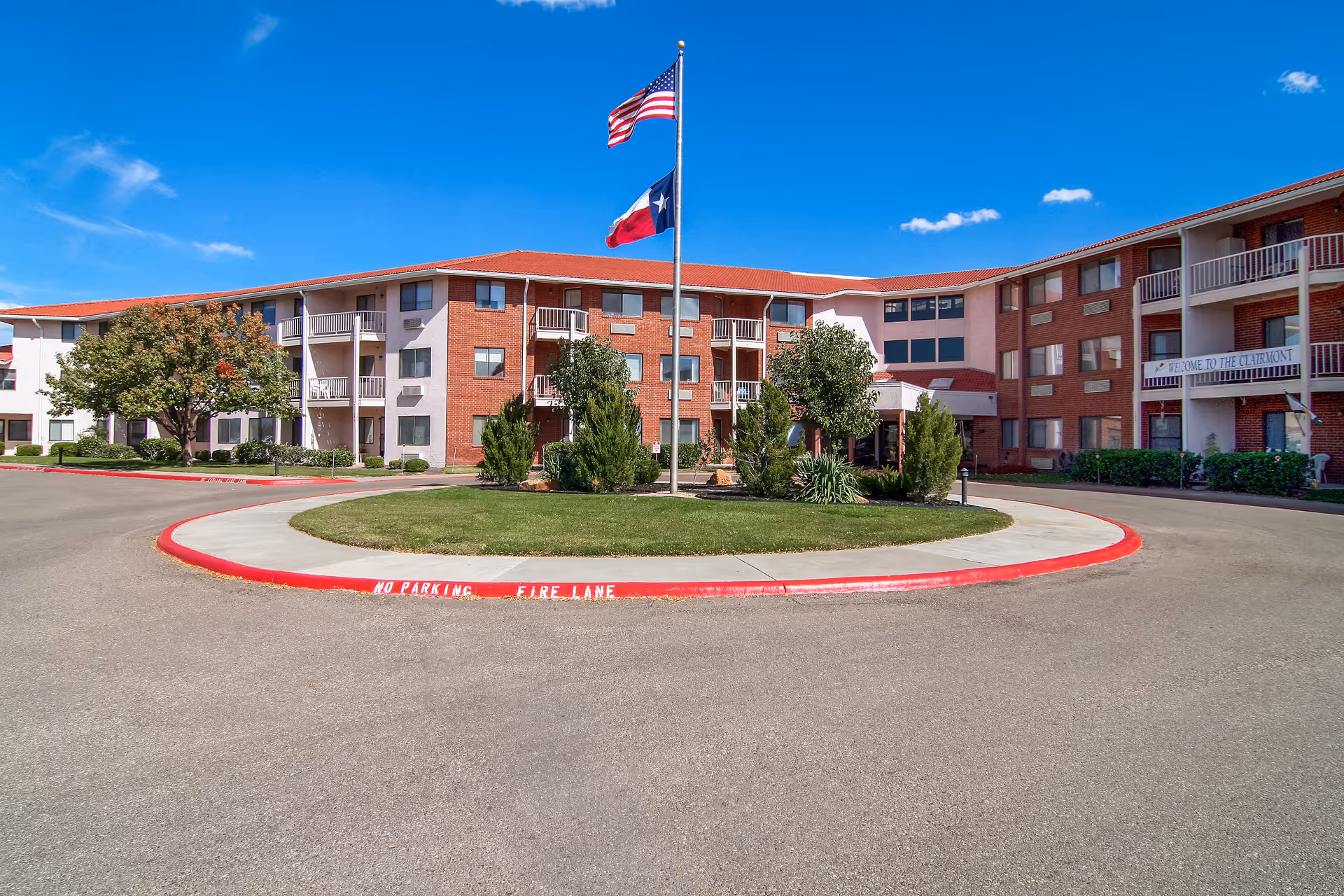 Exterior view of a three-story senior living facility building with red brick and white walls, balconies, and a circular driveway with a grassy island in the center. Two flagpoles display the American and Texas flags under a clear blue sky.
