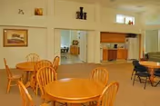A spacious common area with round wooden tables and chairs arranged for seating. The room features a kitchenette with wooden cabinets, a microwave, and a coffee maker. There are large windows allowing natural light to brighten the space, and framed artwork is displayed on the walls.