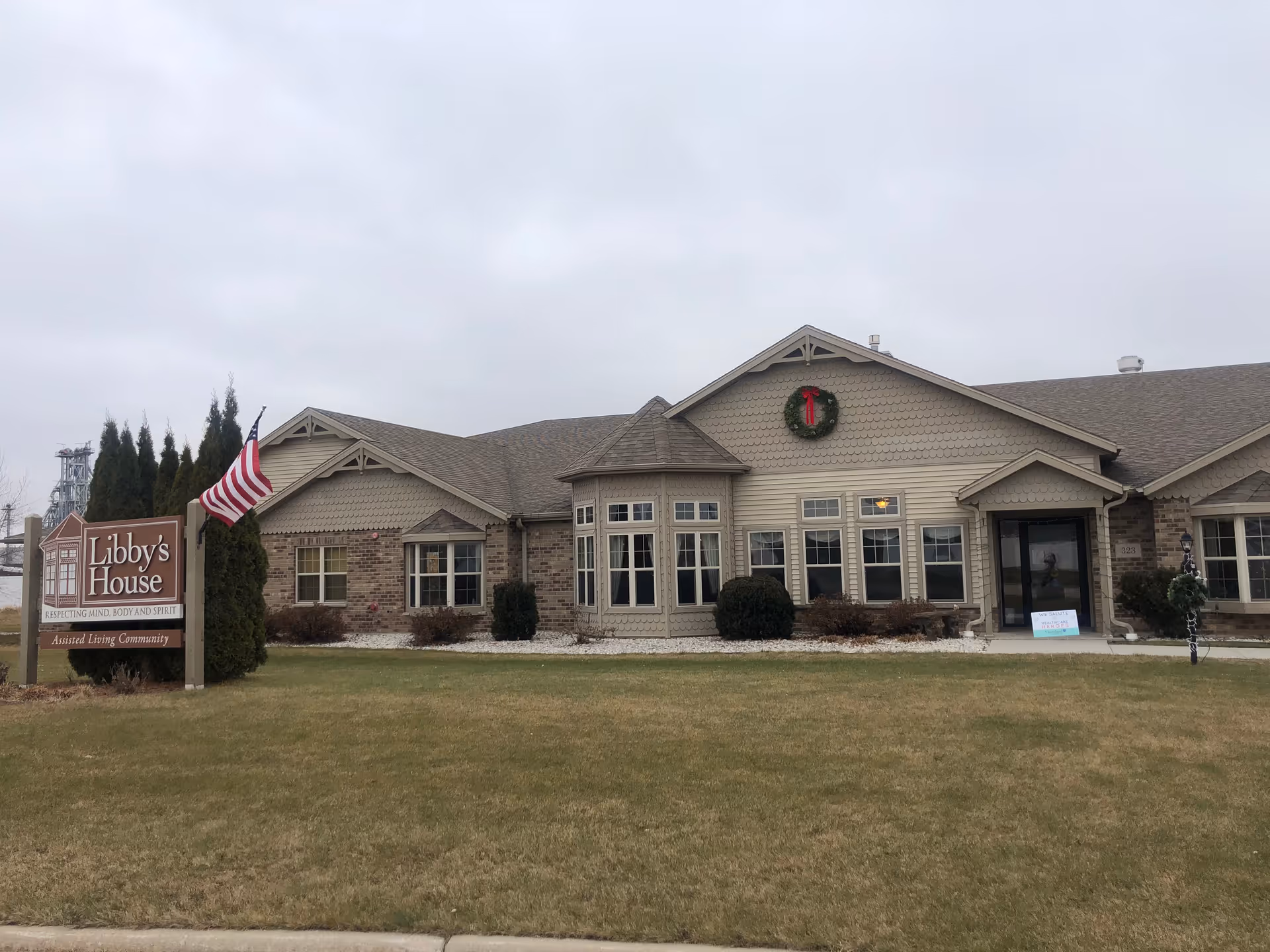 Exterior view of a single-story assisted living community building named Libby's House, with a lawn in front, an American flag on a pole, and a holiday wreath hanging on the building. The sky is overcast.