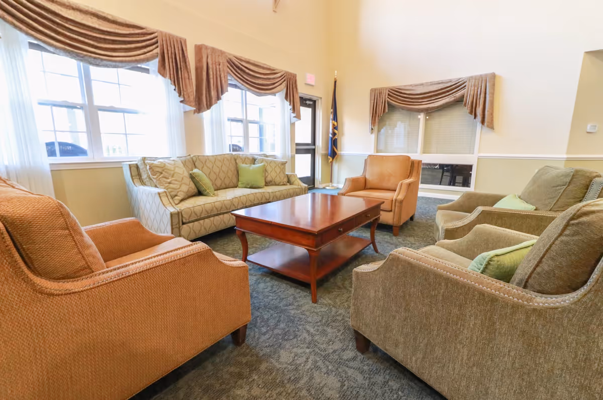 A bright sitting room with sofas, armchairs, and a central wooden coffee table by large windows.