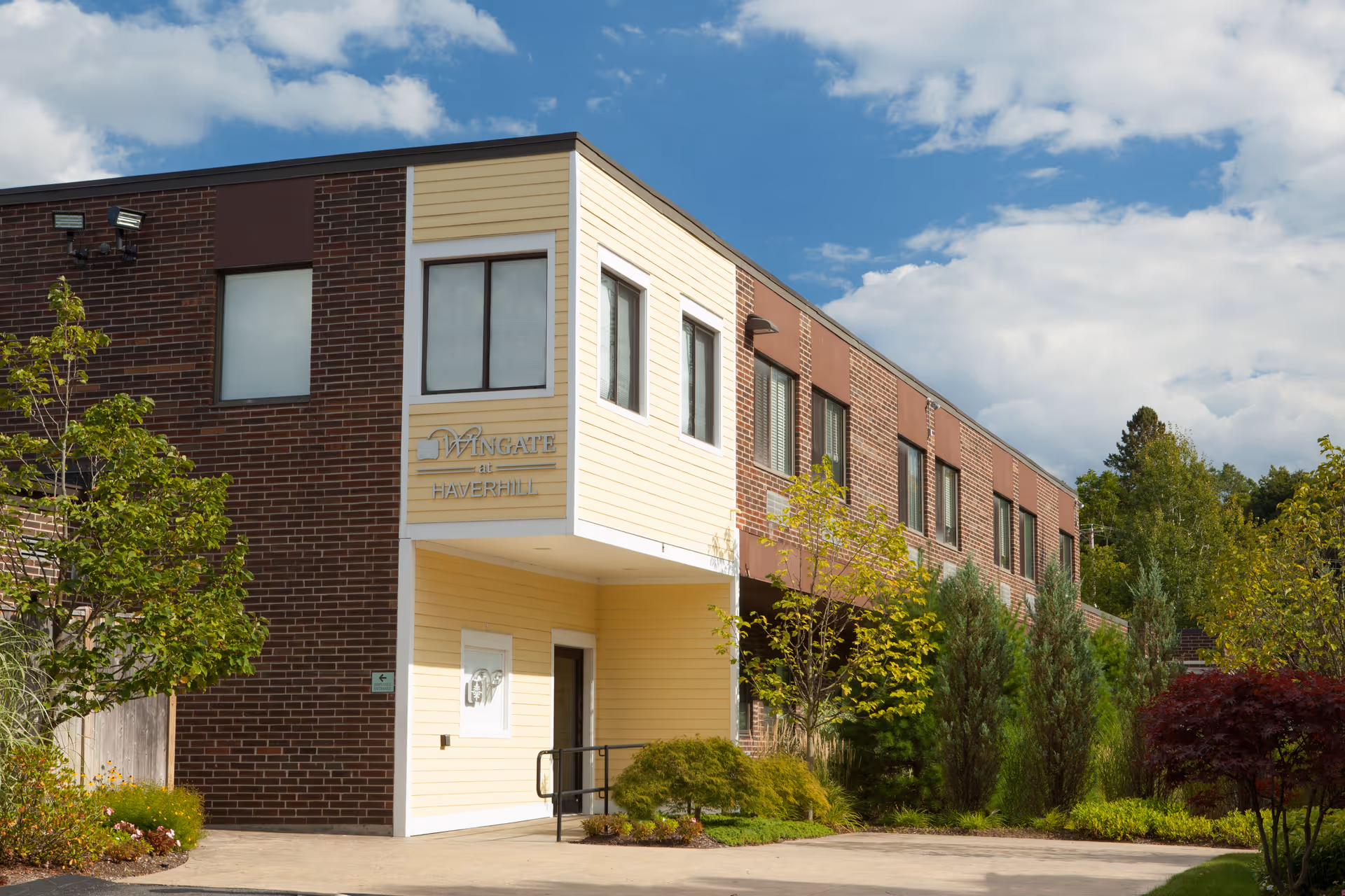 Exterior view of Wingate at Haverhill building featuring a two-story structure with a combination of yellow siding and brown brick walls, several windows, a small entrance with a ramp, and landscaped greenery including trees and shrubs under a partly cloudy sky.