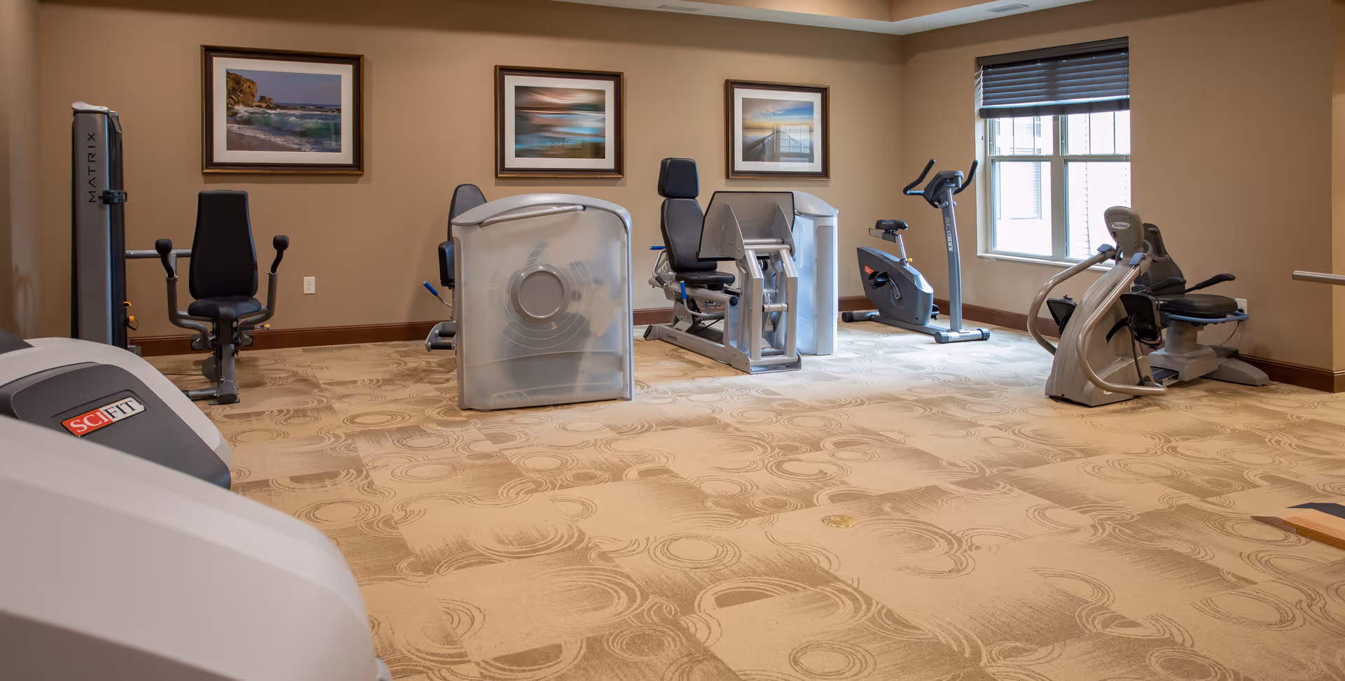 A senior living facility exercise room with various fitness equipment including stationary bikes, a leg press machine, and other workout machines. The room has beige walls, patterned carpet, three framed landscape pictures on the wall, and a window with blinds letting in natural light.