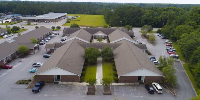 Aerial view of Compass Post Acute Rehabilitation facility showing multiple connected buildings with pitched roofs, surrounded by parking lots and greenery, with a forested area in the background.