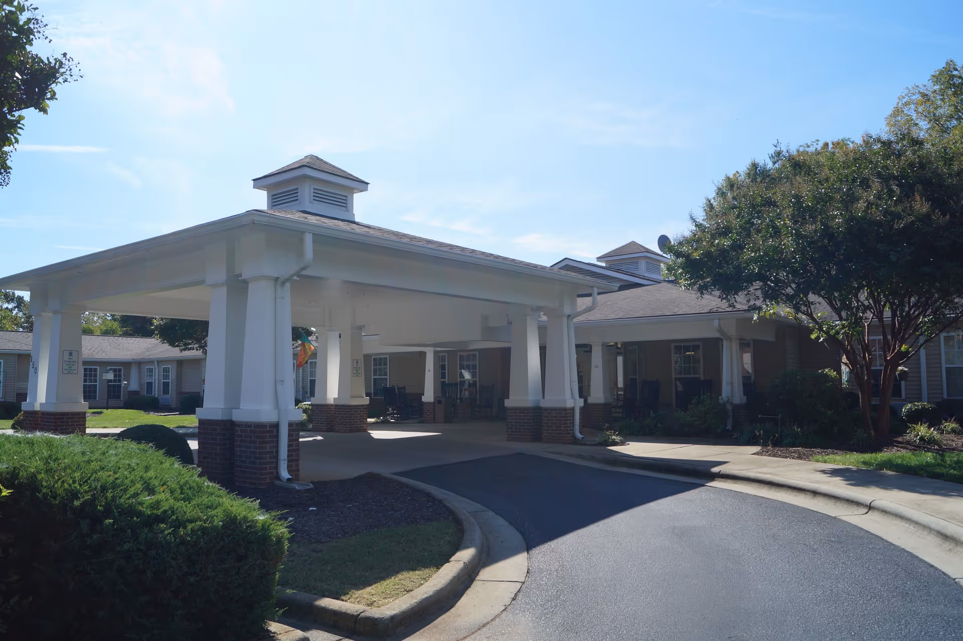 Exterior view of Somerset Court of Mocksville showing a covered driveway entrance with white pillars and brick bases, surrounded by greenery and trees under a clear blue sky.