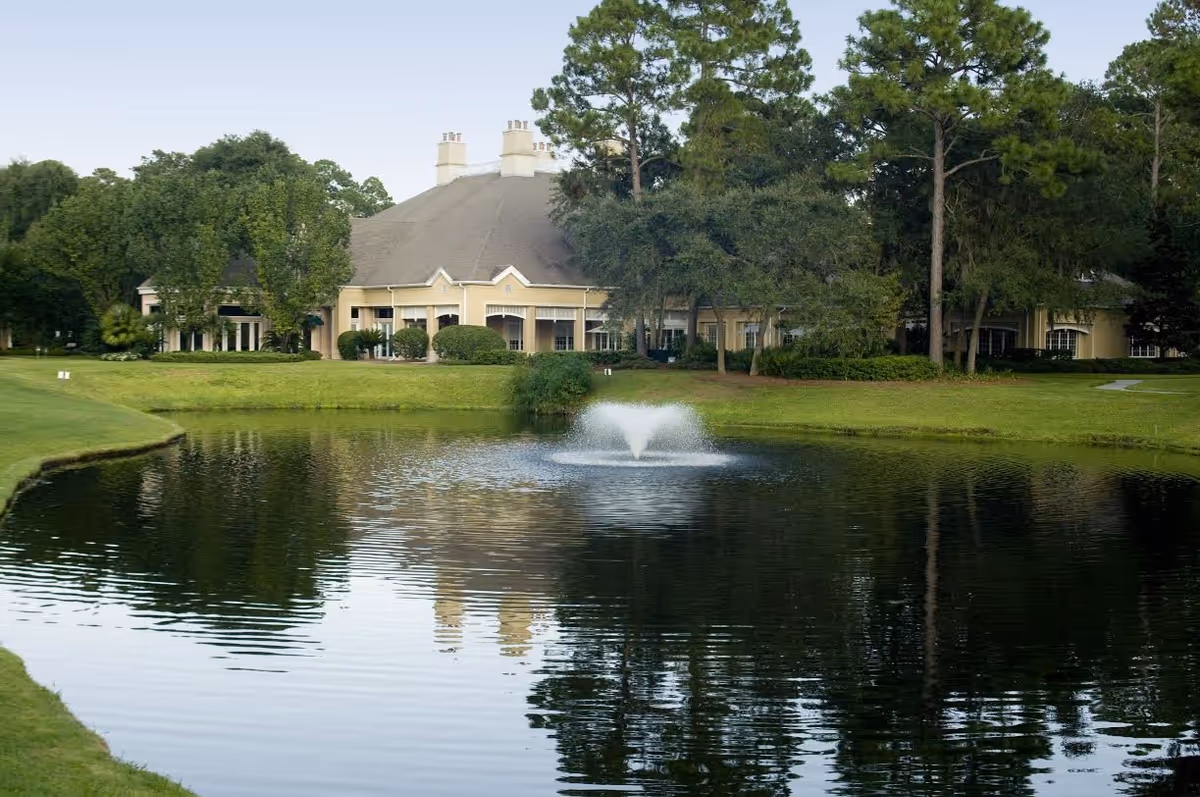 A large building with a steep roof and multiple chimneys is situated behind a pond with a central water fountain. The building is surrounded by green grass, trees, and shrubs, reflecting in the calm water of the pond.