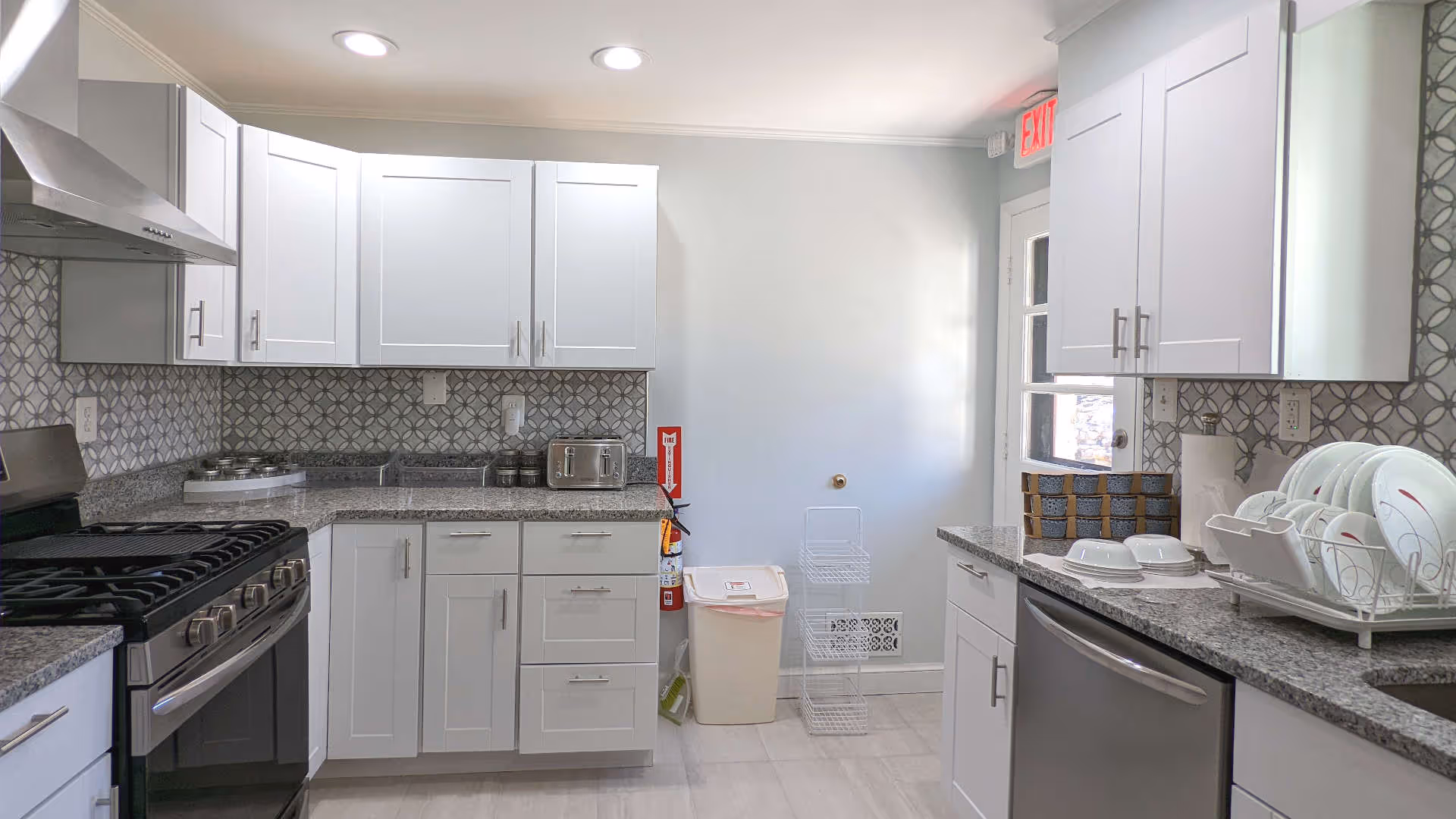A modern kitchen with white cabinets, gray granite countertops, and a patterned tile backsplash. The kitchen features a stainless steel gas stove, a toaster, a dish rack with white dishes, a dishwasher, and a trash bin. There is a door with a window and an exit sign above it.