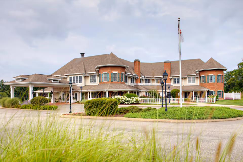 Front exterior of a large two-story brick and siding senior living building with a covered roundabout entrance, flagpole, and landscaped grounds.