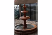 A three-tiered brown stone water fountain in an outdoor area with gravel ground cover and large plants near a building with large glass windows and vertical blinds.