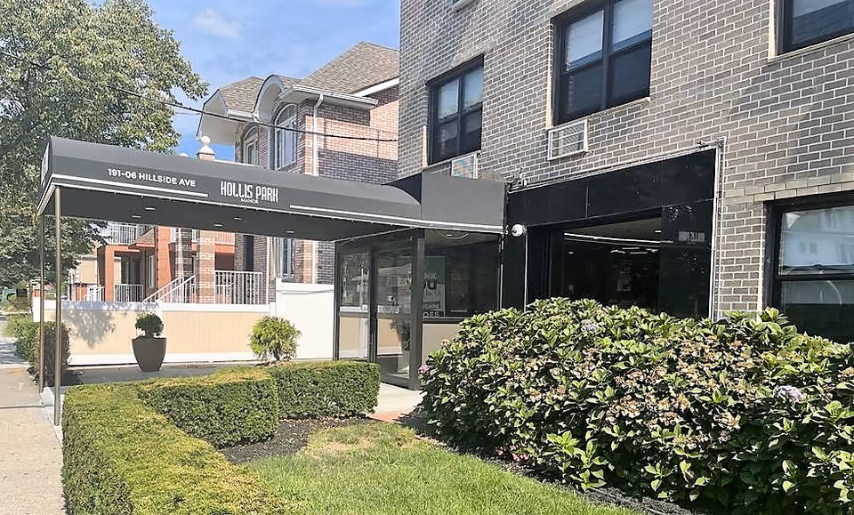 Exterior view of Hollis Park Manor showing the entrance with a black canopy displaying the address 191-06 Hillside Ave and the facility name. The building is made of brick and surrounded by green shrubs and a small lawn area.