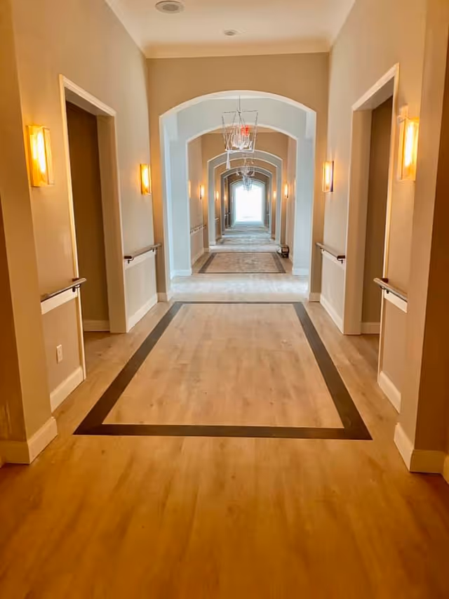Long well-lit interior corridor with repeated archways, chandeliers, wall sconces, wood flooring, and handrails.