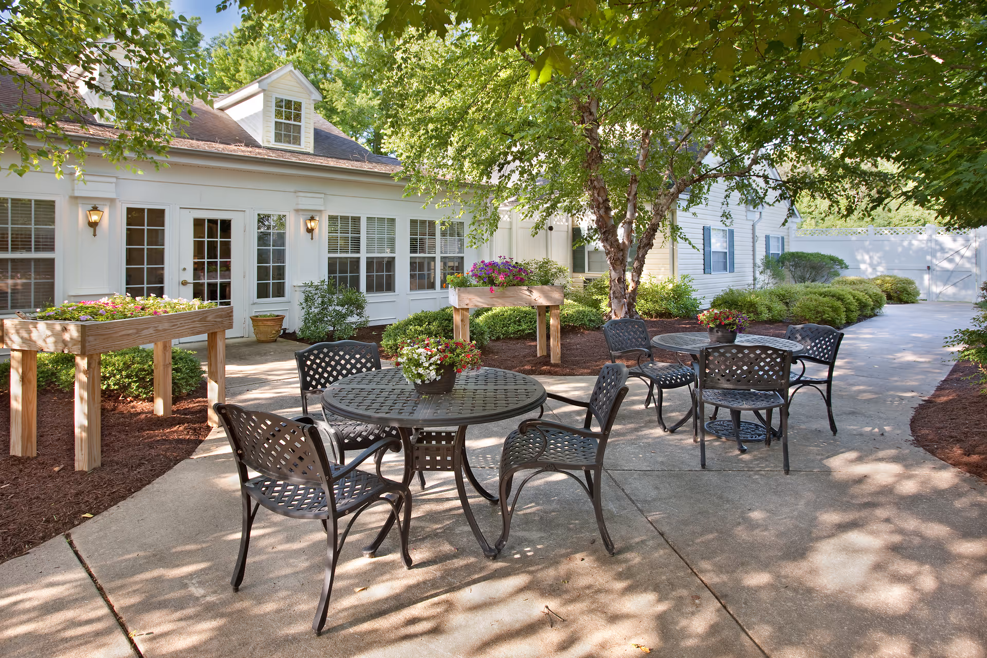 Outdoor patio area at Candle Light Cove featuring metal tables and chairs with flower pots on top, surrounded by greenery and trees, adjacent to a white building with multiple windows and doors.
