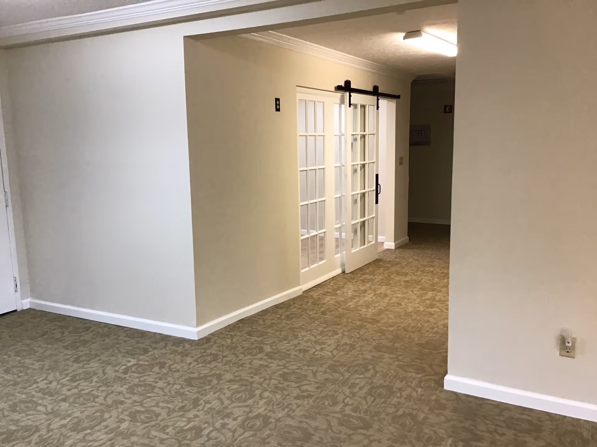 Interior hallway with beige patterned carpet and cream-colored walls. There is a set of white sliding glass doors on a black track leading to another room. The ceiling has crown molding and a fluorescent light fixture.
