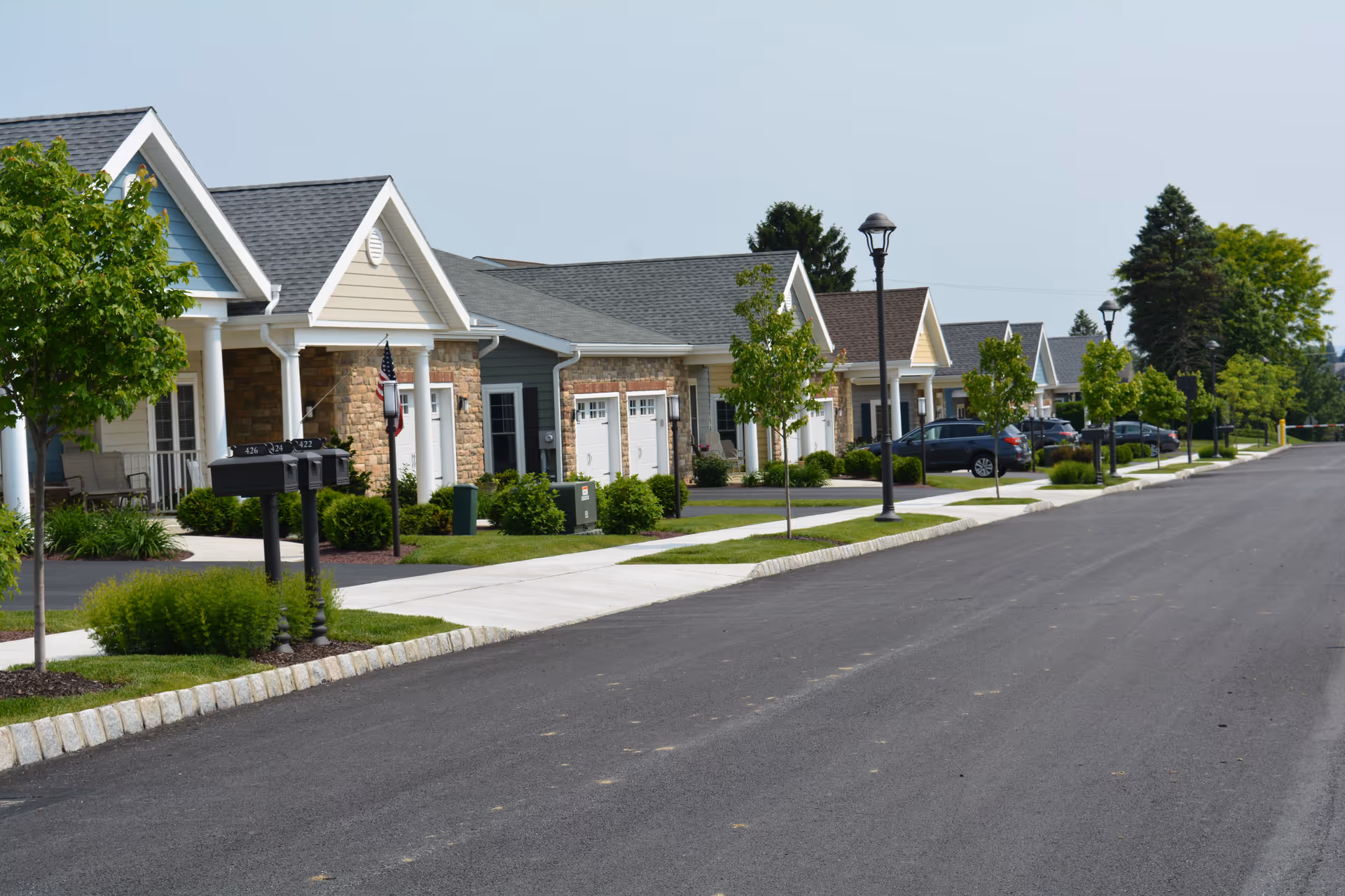 A row of single-story residential buildings with garages, small front porches, and neatly maintained landscaping along a paved street with sidewalks and street lamps under a clear sky.