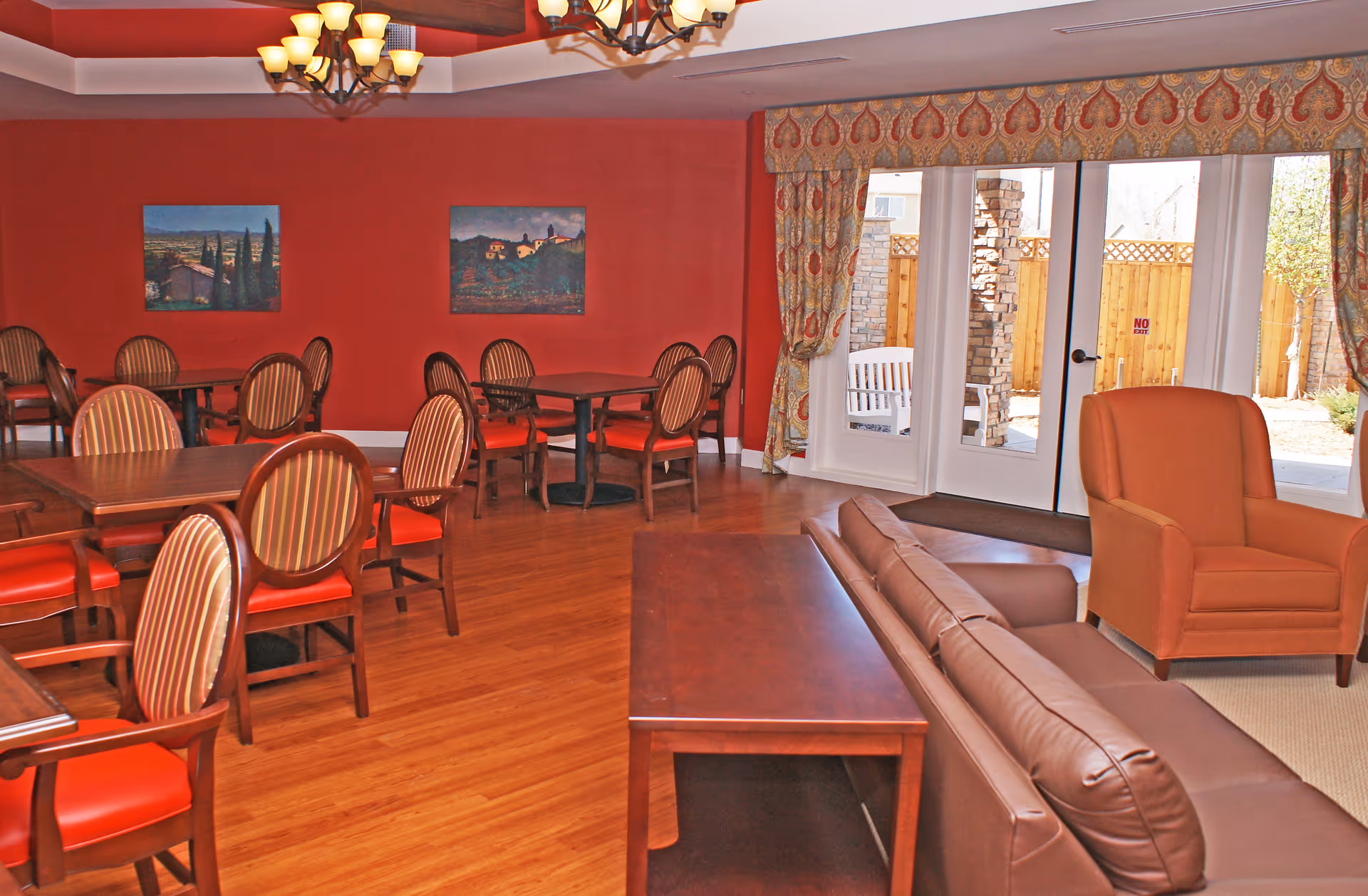 A cozy common area with red walls and wooden flooring featuring several wooden tables with striped cushioned chairs. There is a brown leather sofa and an orange armchair near a set of glass doors that lead outside. The room is decorated with two landscape paintings on the wall and has a chandelier hanging from the ceiling.