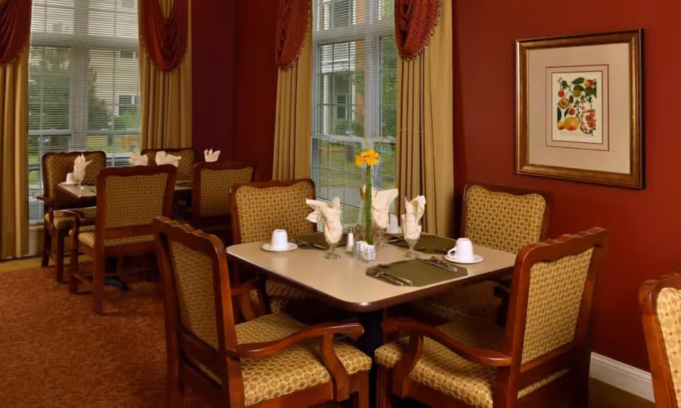 A cozy dining room with several tables and cushioned chairs arranged neatly. Each table is set with folded napkins, cups, glasses, and silverware. The room has large windows with beige curtains and red valances, allowing natural light to brighten the space. A framed botanical print hangs on the red wall.