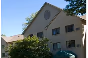 Exterior view of a multi-story residential building with beige siding, several windows, and a triangular roof section. There are trees and greenery in front of the building under a clear blue sky.