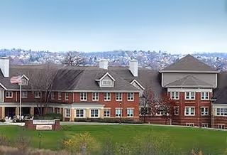 Exterior view of a large multi-story senior living facility building with red brick and beige siding, multiple windows, and a well-maintained green lawn in the foreground under a clear sky.