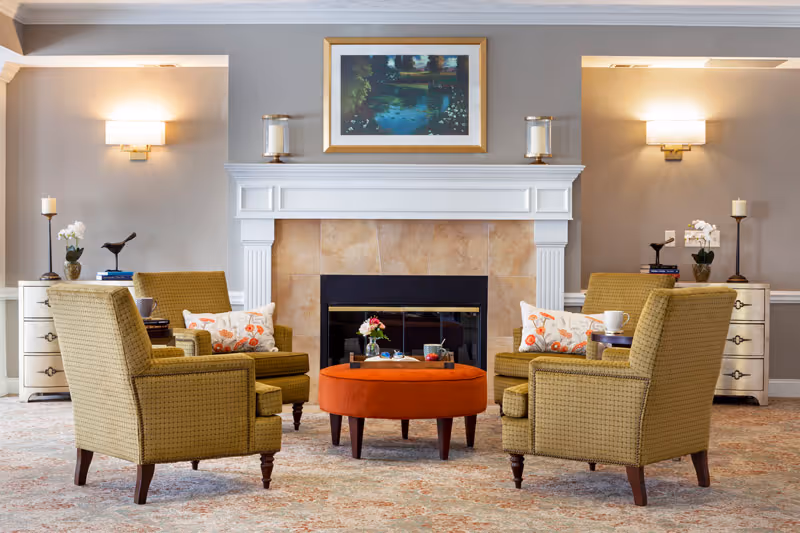 Cozy seating area with four upholstered armchairs arranged around an orange ottoman in front of a fireplace topped by framed artwork.